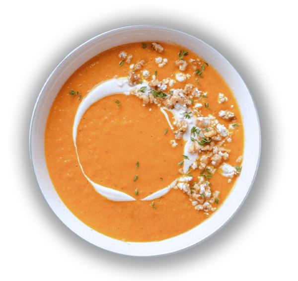 A bowl of creamy orange soup garnished with a swirl of cream, crumbled pieces, and fresh herbs. The soup sits on a gray surface, creating a warm and inviting presentation. GREENHOUSE Goodness A bowl of creamy orange soup garnished with a swirl of cream, crumbled pieces, and fresh herbs. The soup sits on a gray surface, creating a warm and inviting presentation. GREENHOUSE Goodness