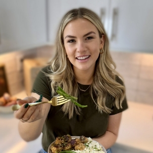 A person with long blonde hair, wearing a dark green shirt, is smiling and holding a fork with green beans over a plate of food in a kitchen setting. GREENHOUSE Goodness A person with long blonde hair, wearing a dark green shirt, is smiling and holding a fork with green beans over a plate of food in a kitchen setting. GREENHOUSE Goodness