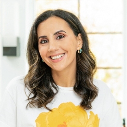 A person with long, wavy dark hair smiles warmly at the camera. They are wearing a white shirt with a yellow floral design and small hoop earrings. A softly blurred background suggests a well-lit room with large windows. GREENHOUSE Goodness A person with long, wavy dark hair smiles warmly at the camera. They are wearing a white shirt with a yellow floral design and small hoop earrings. A softly blurred background suggests a well-lit room with large windows. GREENHOUSE Goodness