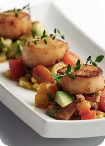 Three seared scallops garnished with herbs on a rectangular white plate. They are served over a colorful bed of diced avocado, tomatoes, and other vegetables, with a light sauce drizzle. GREENHOUSE Goodness Three seared scallops garnished with herbs on a rectangular white plate. They are served over a colorful bed of diced avocado, tomatoes, and other vegetables, with a light sauce drizzle. GREENHOUSE Goodness