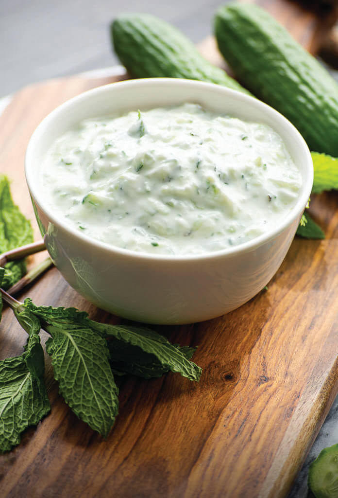 Bowl of tzatziki sauce with visible cucumber and herbs on a wooden board. Mint leaves and cucumbers are nearby. GREENHOUSE Goodness