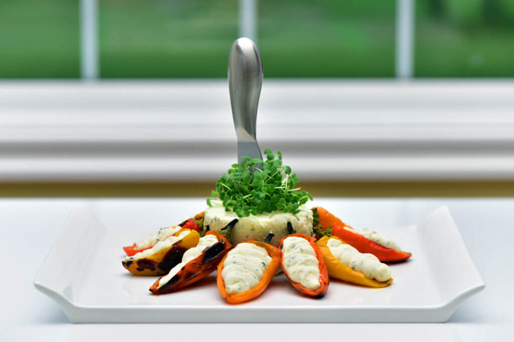 A dish on a white plate features stuffed peppers with a creamy filling, arranged around a mound of greens. A fork is vertically stabbed into the greens. The background shows a blurred window view of greenery. GREENHOUSE Goodness