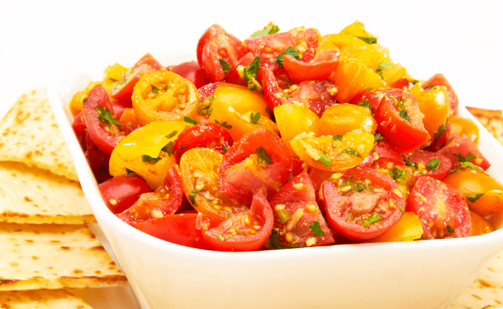 A white bowl filled with a colorful mix of cherry tomatoes in red and yellow, garnished with herbs. The background includes stacked, triangular tortilla chips. The vibrant colors of the salad contrast with the neutral background. GREENHOUSE Goodness