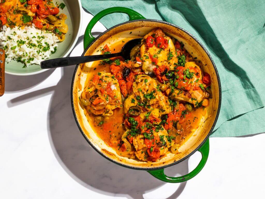 A green pot filled with chicken thighs cooked with cherry tomatoes, herbs, and vegetables. A black spoon is placed inside the pot. A bowl of white rice and vegetables is partially visible nearby, with a green cloth beside it. GREENHOUSE Goodness