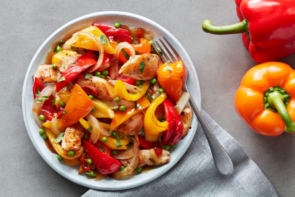 A colorful dish featuring sautéed chicken pieces mixed with red, yellow, and orange bell peppers, onions, and peas on a white plate. A fork rests on the plate, next to fresh red and orange bell peppers, atop a gray napkin on a table. GREENHOUSE Goodness