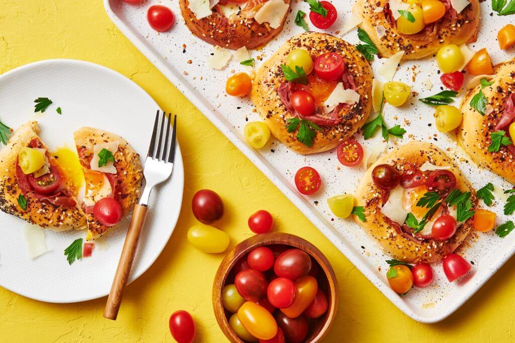 Bagel pizzas topped with sliced cherry tomatoes, herbs, and cheese on a tray and a plate, surrounded by a bowl of tomatoes on a vibrant yellow background. GREENHOUSE Goodness
