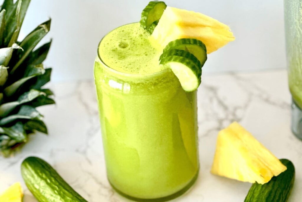 A frothy Cucumber Pineapple Smoothie in a glass, garnished with greenhouse mini cucumber slices and pineapple chunks. Fresh cucumber and pineapple pieces are scattered on a marble surface nearby. A whole pineapple is partially visible in the background. GREENHOUSE Goodness