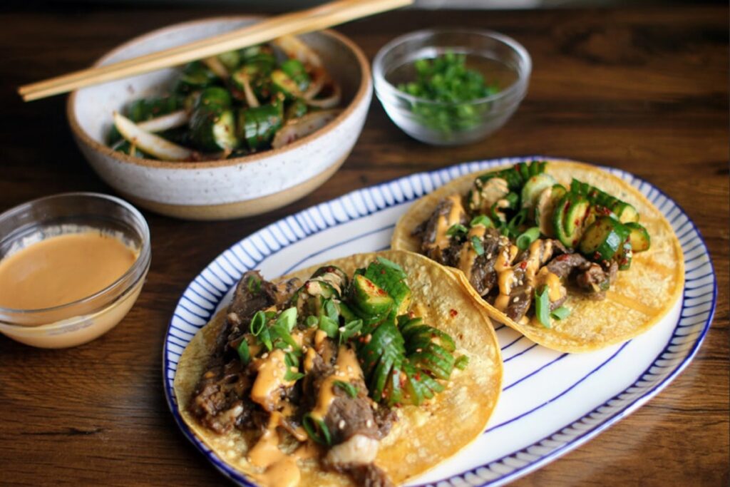 Two bulgogi tacos filled with sliced beef, green onions, and cucumbers drizzled with a creamy sauce on a striped plate. In the background, a bowl of sliced cucumber salad with onions, alongside small bowls of green onions and sauce. GREENHOUSE Goodness