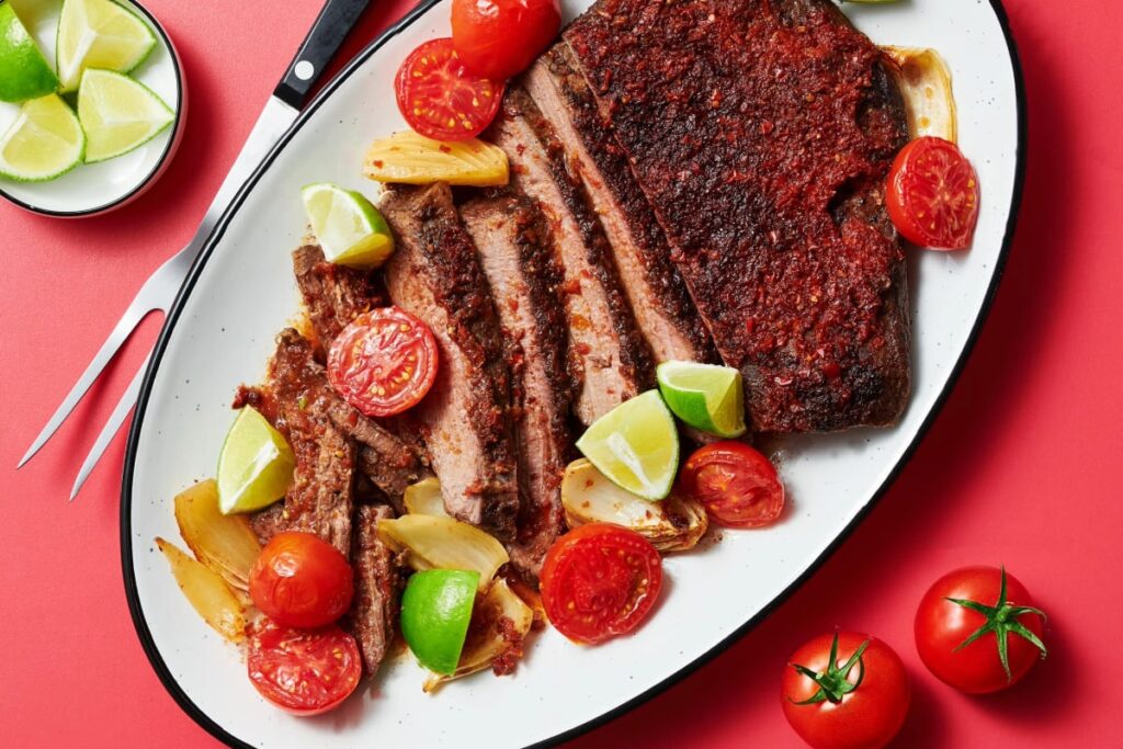 A plate of savory, seasoned beef brisket with grilled onions complements seared scallops paired beautifully with a corn, avocado, and bacon salsa. Quartered limes and halved cherry tomatoes add freshness. A carving fork rests beside the dish on a vibrant red background. GREENHOUSE Goodness