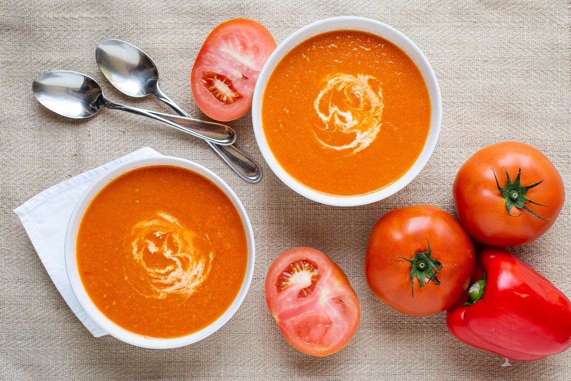 Two bowls of Creamy Tomato Basil & Parmesan Soup with a swirl of cream rest on a beige cloth, surrounded by fresh tomatoes, a sliced tomato, red bell pepper, and two spoons. GREENHOUSE Goodness