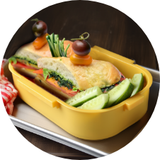 A yellow lunchbox containing a sandwich with lettuce, tomato, and meat, garnished with cherry tomatoes on skewers. Sliced cucumbers are placed alongside the sandwich. The lunchbox rests on a metal tray. GREENHOUSE Goodness