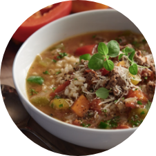A bowl of hearty soup containing minced meat, rice, diced tomatoes, and vegetables, topped with grated cheese and fresh herbs. The soup is served in a white bowl on a wooden table, with a spoon nearby. GREENHOUSE Goodness