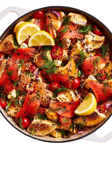 A colorful dish with smoked salmon, bagel pieces, cherry tomatoes, lemon wedges, dill, and sesame seeds in a white pan. GREENHOUSE Goodness