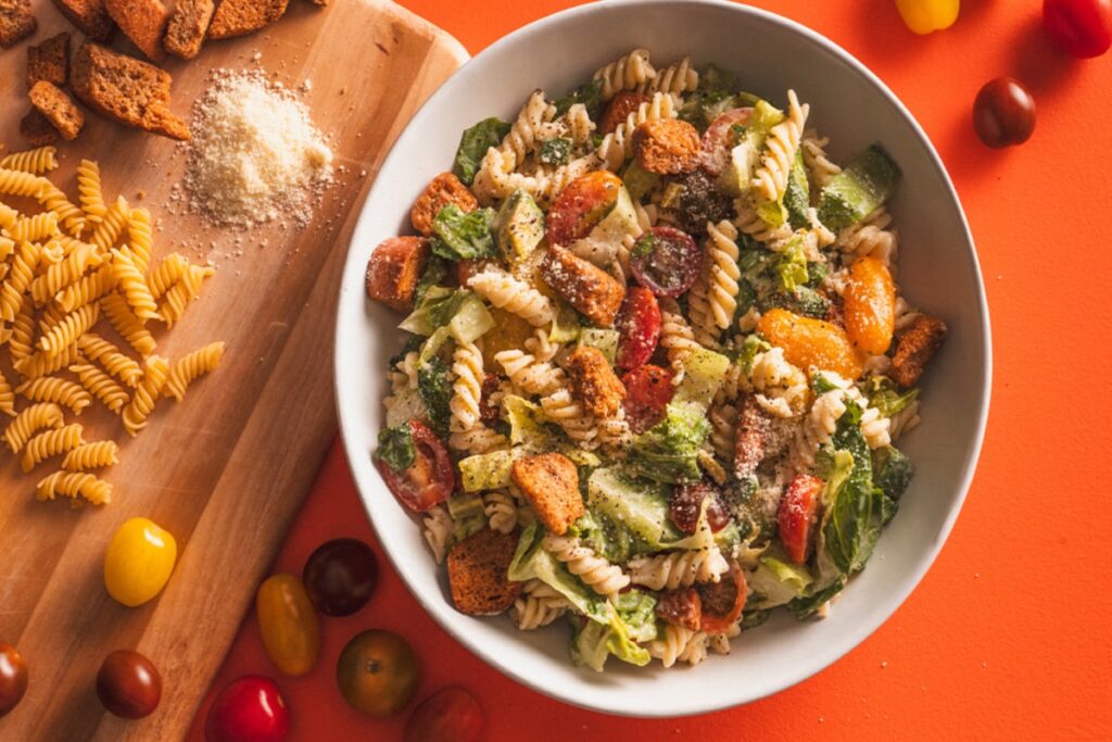 A vibrant Caesar pasta salad featuring rotini, crisp lettuce, cherry tomatoes, crunchy croutons, and grated cheese is set against an orange backdrop. Beside it, a wooden board displays raw pasta and a pile of cheese, surrounded by assorted cherry tomatoes. GREENHOUSE Goodness