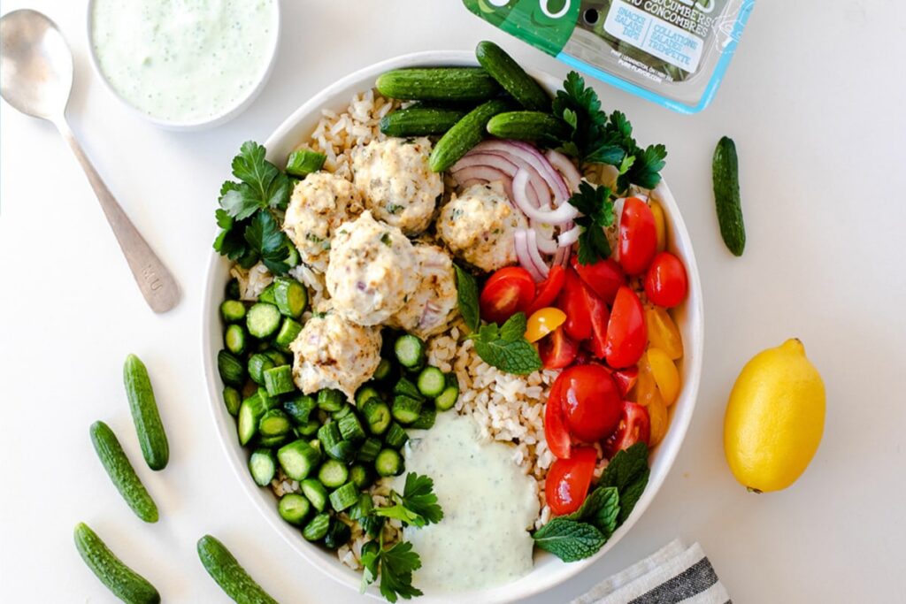 A vibrant Greek Chicken Meatball Bowl brimming with flavor: rice crowned with succulent chicken meatballs, sliced cucumbers, cherry tomatoes, red onions, and aromatic herbs. Accompanied by a zesty green sauce, a fresh lemon wedge, and a spoon on a pristine white surface. GREENHOUSE Goodness