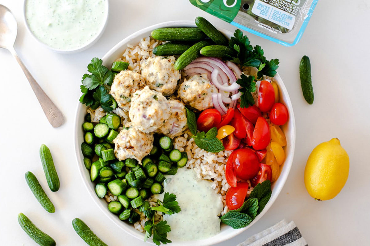 A Greek Chicken Meatball Bowl featuring rice, chicken meatballs, sliced cucumbers, cherry tomatoes, red onion, parsley, mint, and yogurt sauce sits on a white surface with mini cucumbers and a lemon nearby. GREENHOUSE Goodness