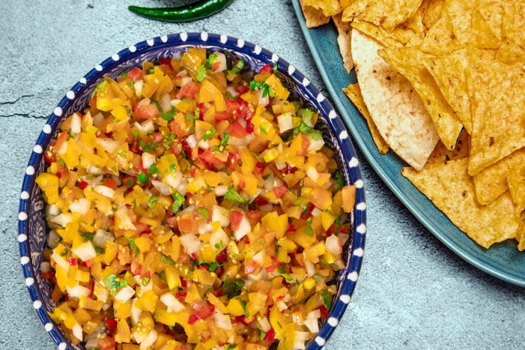 A bowl of vibrant homemade salsa with chopped yellow and red tomatoes, onions, and herbs sits on a blue speckled plate. Next to it, a platter holds a stack of golden tortilla chips. Two green chili peppers rest nearby on a gray surface. GREENHOUSE Goodness