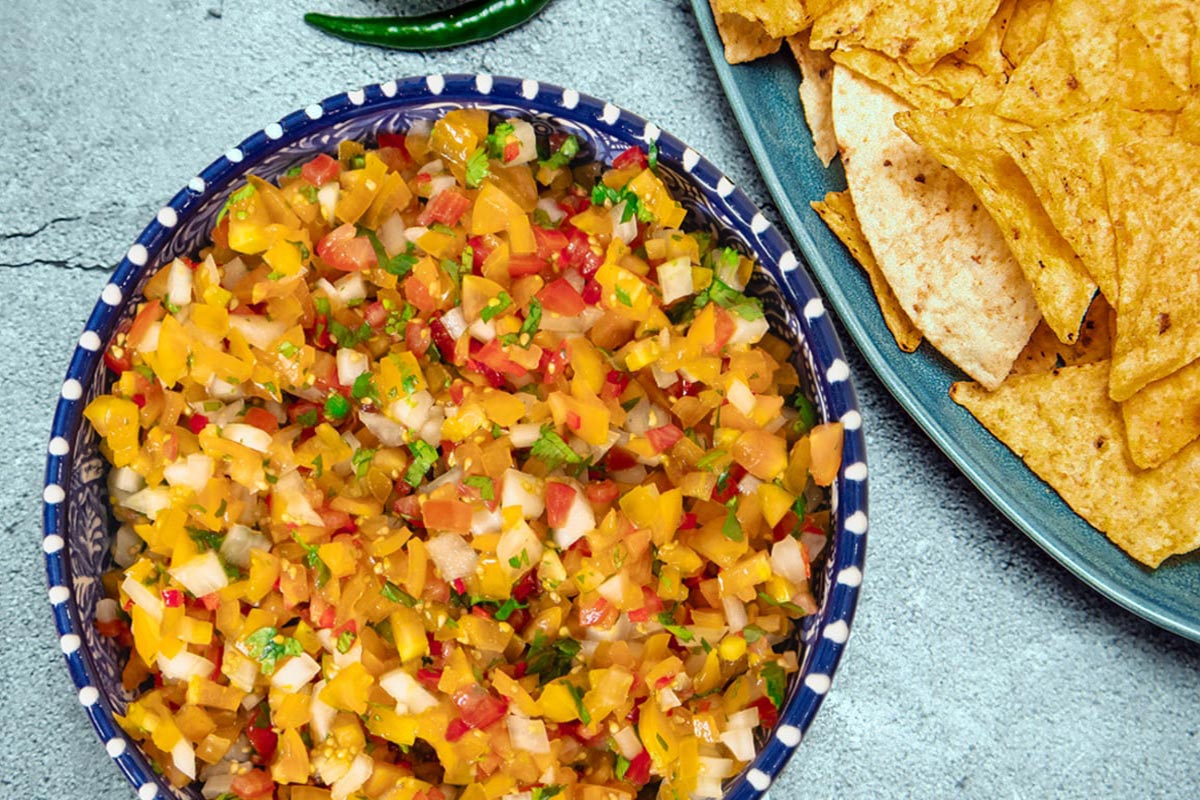 A bowl of colorful homemade salsa made with diced tomatoes, onions, and peppers sits next to a plate of tortilla chips on a textured surface. Two green chili peppers are also visible at the top of the image. GREENHOUSE Goodness