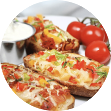 Close-up of three slices of toasted bread topped with melted cheese, chopped tomatoes, bell peppers, and herbs on a white plate. Fresh cherry tomatoes and a small cup of creamy dip are in the background. GREENHOUSE Goodness