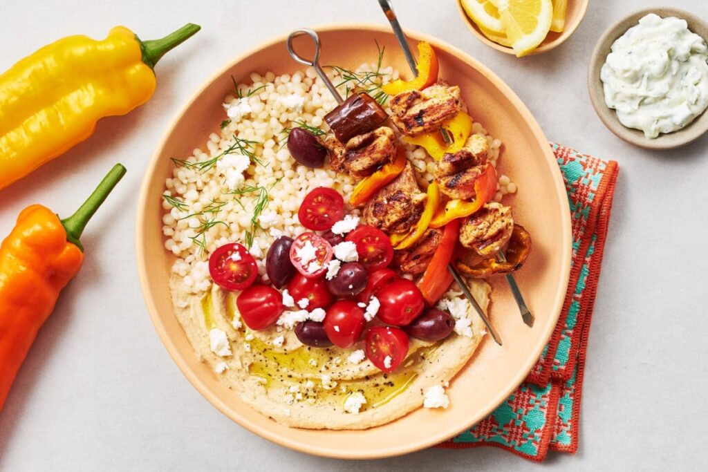 A vibrant Grilled Shawarma Mezze Bowl features a bed of couscous, richly hummus-drizzled with olive oil, cherry tomatoes, and olives. Topped with skewers of grilled chicken and bell peppers, it comes with a side of tzatziki and lemon wedges, surrounded by colorful yellow and orange peppers. GREENHOUSE Goodness