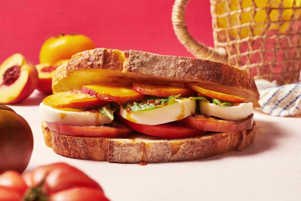 A Peach Caprese Sandwich with heirloom tomatoes, fresh mozzarella, and greens is placed on a light surface. Whole tomatoes and peaches lie in the background, accompanied by a netted basket and a pink backdrop. GREENHOUSE Goodness