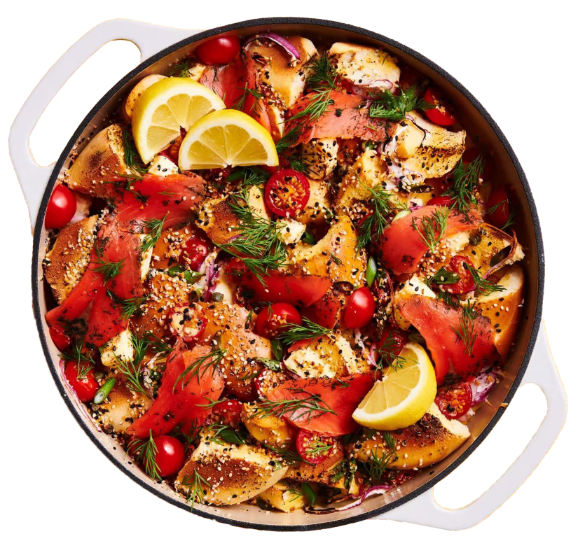 A round baking dish with white handles filled with bagel pieces, smoked salmon, cherry tomatoes, and fresh dill. Lemon slices are garnished on top, and everything is sprinkled with seasoning. GREENHOUSE Goodness