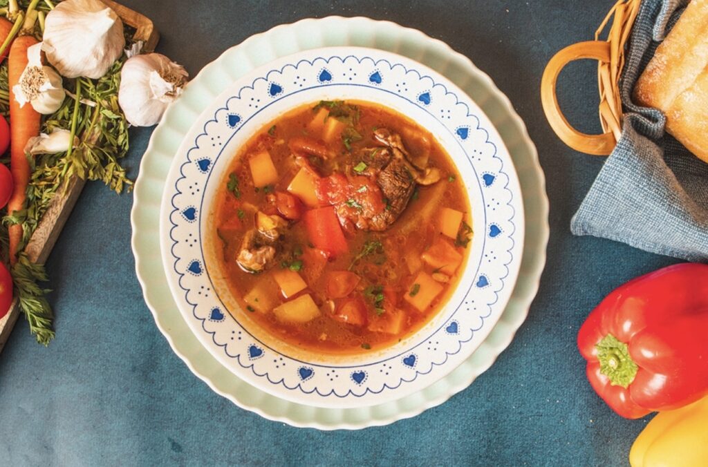 A bowl of hearty braised oxtail and vegetable stew garnished with herbs. The soup contains chunks of beef, potatoes, and tomatoes. Surrounding the bowl are garlic, a red bell pepper, a loaf of bread, and a basket on a blue surface. GREENHOUSE Goodness