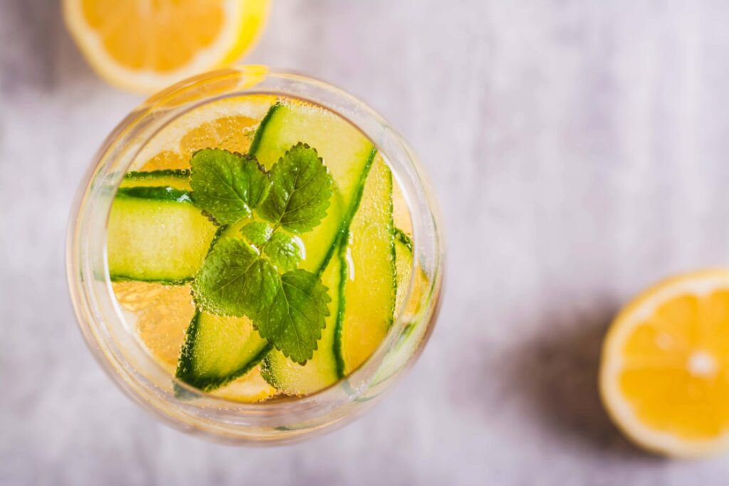 Top view of a refreshing cucumber melon sangria mocktail with cucumber slices and mint leaves in a glass. Lemon slices are on the side on a gray marble surface. GREENHOUSE Goodness