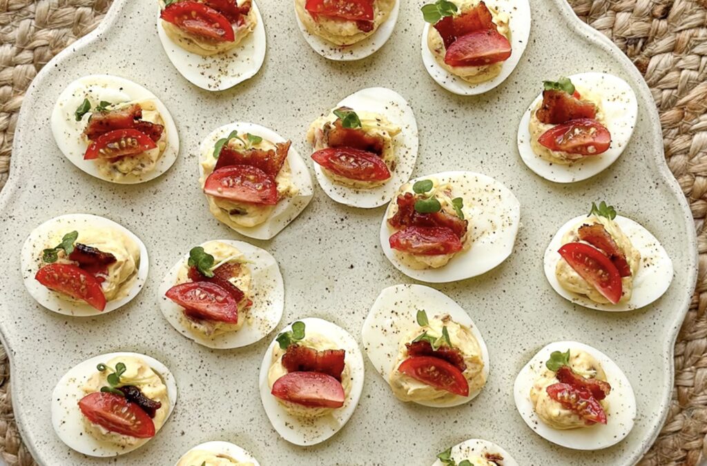 Plate of deluxe BLT deviled eggs topped with sliced cherry tomatoes and herbs on a textured wicker surface. The eggs are neatly arranged in a circular pattern. GREENHOUSE Goodness