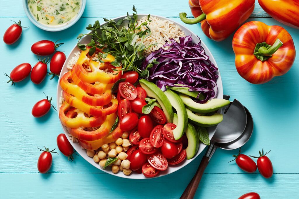 A vibrant Rainbow Buddha Bowl with sliced bell peppers, cherry tomatoes, cucumber, purple cabbage, chickpeas, and rice on a turquoise background. Whole bell peppers and cherry tomatoes are scattered around, with dressing on the side. GREENHOUSE Goodness