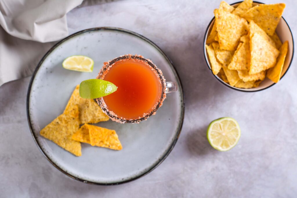 A savory, red brunch mocktail in a glass with a salted rim and lime garnish is on a plate with tortilla chips. Nearby, a bowl of tortilla chips and a lime wedge rest on a light gray surface. GREENHOUSE Goodness