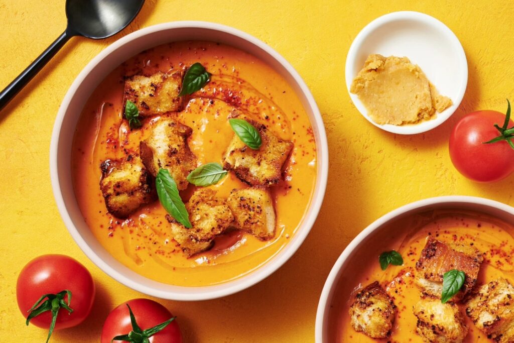 Bowls of creamy tomato soup garnished with croutons, basil, and chili flakes rest on a yellow surface. Nearby, a small dish of hummus and fresh tomatoes accompany the spread, while a black spoon lies in the top left corner. Perfectly paired with The Bella Pepper Mocktail for a refreshing touch. GREENHOUSE Goodness