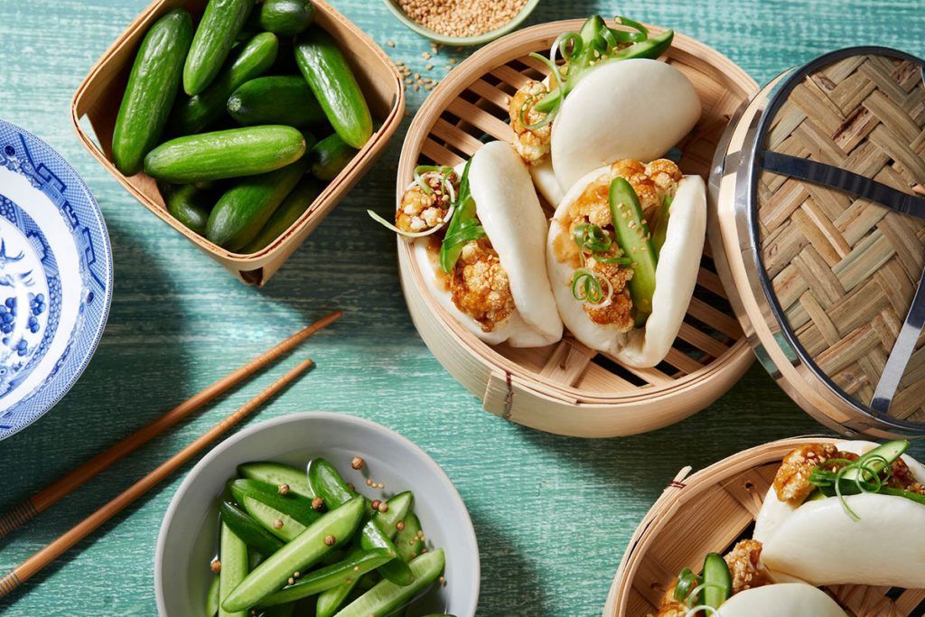 Teriyaki Bao Buns filled with fried cauliflower, sliced cucumber, and green onion in a bamboo steamer, served with snap peas, mini cucumbers, and chopsticks on a green wooden table. GREENHOUSE Goodness