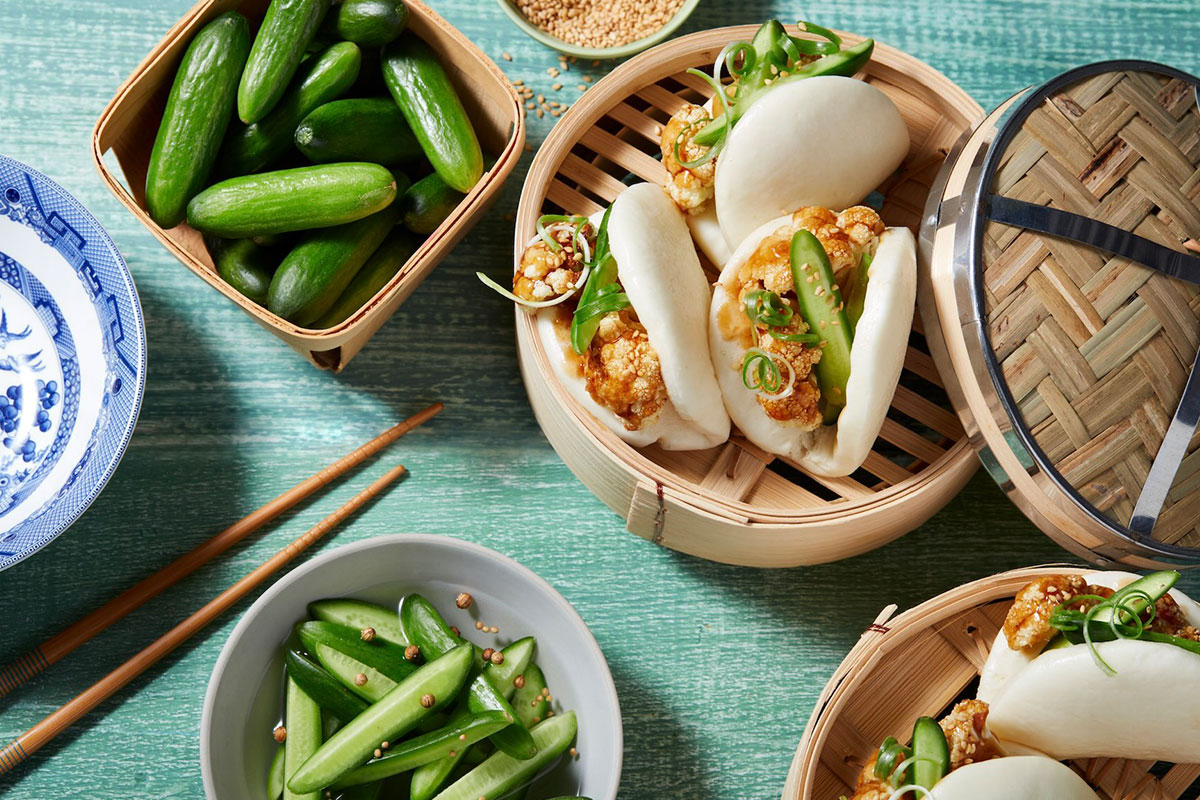 Teriyaki Bao Buns filled with fried cauliflower, sliced cucumber, and green onion in a bamboo steamer, served with snap peas, mini cucumbers, and chopsticks on a green wooden table. GREENHOUSE Goodness