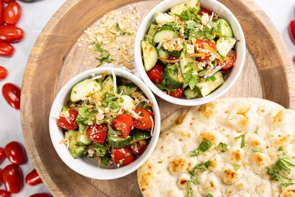 Two bowls of cucumber and tomato salad, resembling a crispy Shred Coco Salad, are garnished with herbs and seeds on a wooden platter. Nearby, a piece of naan sprinkled with herbs complements the display. Cherry tomatoes are scattered around, enhancing the fresh presentation. GREENHOUSE Goodness