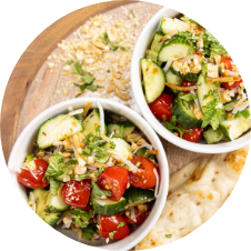 Two bowls of fresh salad with cucumber, cherry tomatoes, herbs, and chopped nuts on a wooden plate. A piece of flatbread is placed beside the bowls. The salad appears vibrant and colorful. GREENHOUSE Goodness