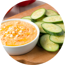 A bowl of creamy hummus topped with a drizzle of oil and spices is set on a wooden board. Next to it are sliced cucumber pieces, perfect for dipping. GREENHOUSE Goodness
