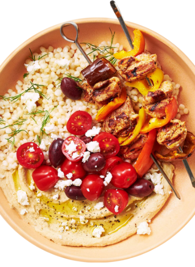 A plate with grilled chicken and vegetable skewers, atop a bed of couscous. Accompanied by cherry tomatoes, black olives, feta cheese, and hummus drizzled with olive oil and sprinkled with seasoning. Garnished with fresh dill. GREENHOUSE Goodness