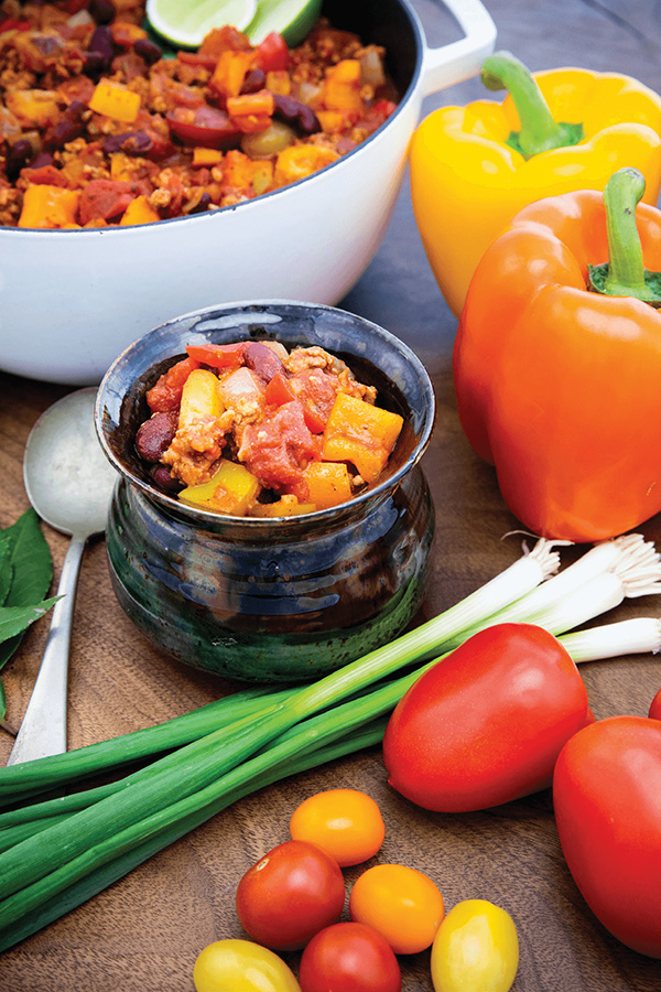 A ceramic bowl filled with chili sits on a wooden surface. Surrounding it are yellow and orange bell peppers, green onions, and assorted tomatoes. A larger pot of chili and a spoon are in the background. GREENHOUSE Goodness