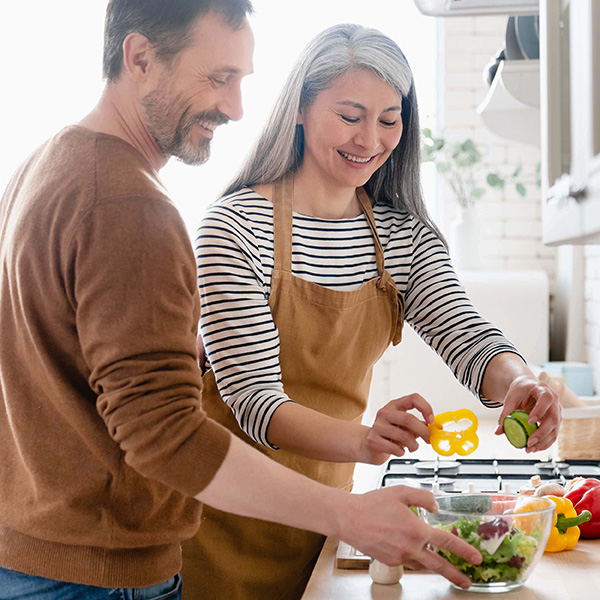 A man and a woman smile while preparing a salad in a bright kitchen. The woman places a yellow bell pepper slice into a bowl, surrounded by various fresh vegetables on the counter. Both are wearing casual clothes, and the woman is in an apron. GREENHOUSE Goodness