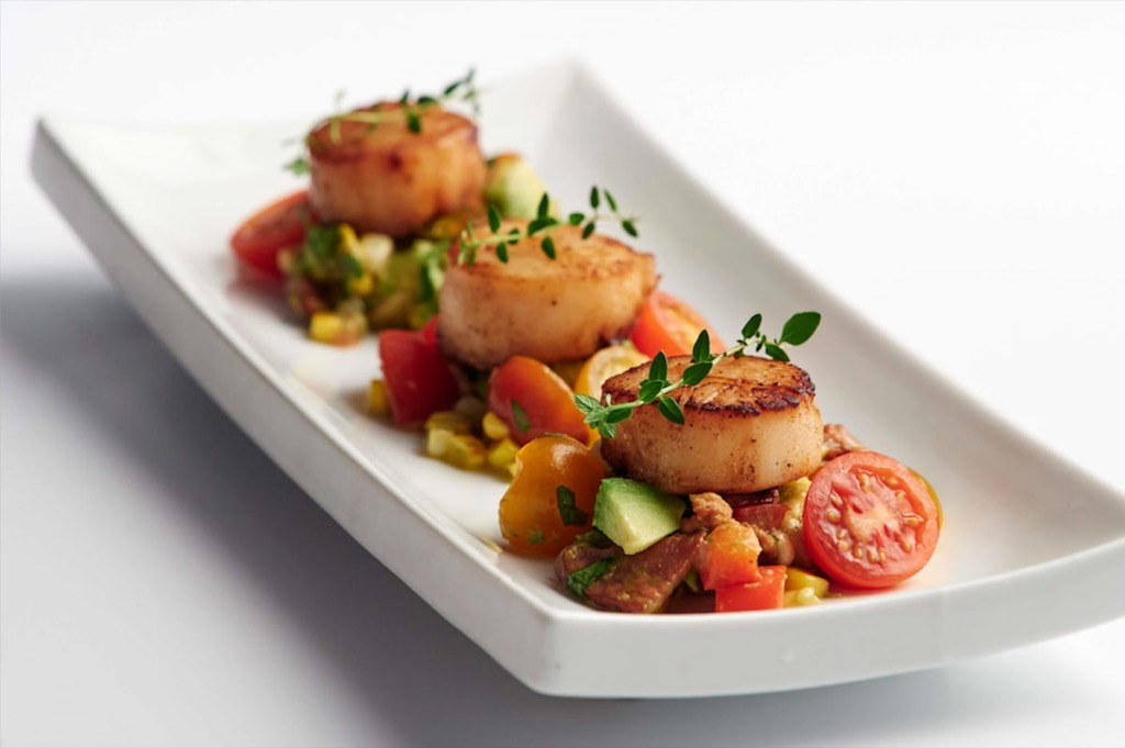 Three seared scallops are served on a rectangular white plate, each atop a colorful medley of diced vegetables and halved Cocktail Tomatoes, garnished with fresh herbs. GREENHOUSE Goodness