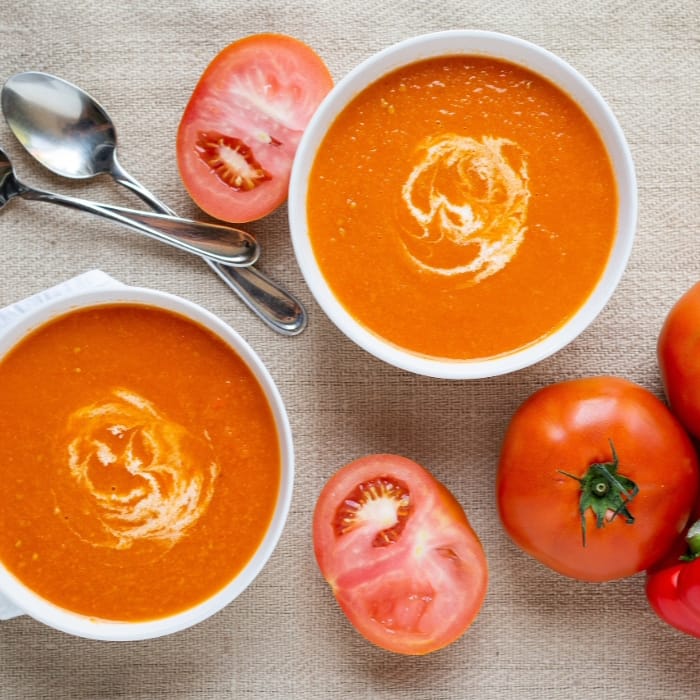 Two bowls of creamy tomato soup with a swirl of cream on top, placed on a textured surface. Fresh tomatoes, some halved, are beside the bowls. Two spoons are on the side. The arrangement creates a warm and appetizing presentation. GREENHOUSE Goodness