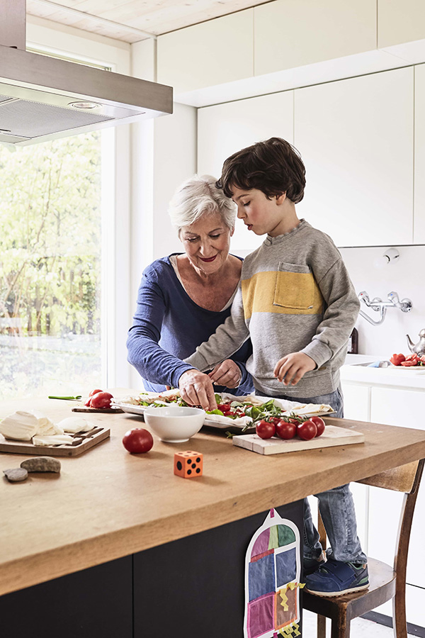 Grandmother and grandson cooking together in a bright kitchen. The woman is wearing a blue top, assisting the boy, who stands on a stool, wearing a gray sweater with a yellow pocket. Fresh ingredients like tomatoes and lettuce are on the countertop. GREENHOUSE Goodness