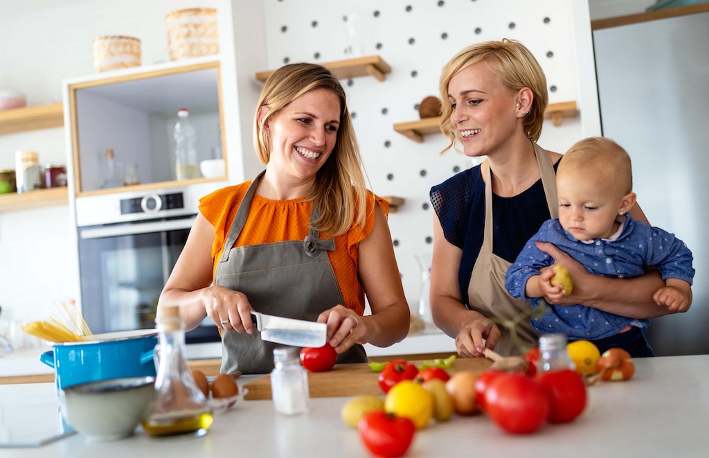 Two women in aprons smile while preparing food in a modern kitchen. One is slicing a Cocktail Tomtato, and the other holds a baby. A variety of fresh produce is spread across the counter, adding color and vibrancy to their culinary endeavor. GREENHOUSE Goodness