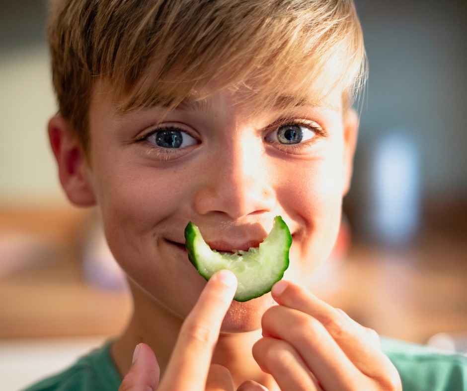 A young boy with blond hair holds a cucumber slice in front of his mouth, making it look like a smile. He has bright blue eyes and is looking directly at the camera with a cheerful expression. The background is softly blurred. GREENHOUSE Goodness