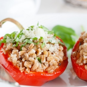 A halved red bell pepper stuffed with cooked grains, garnished with chopped herbs and grated cheese, is placed on a white plate. A green leaf is blurred in the background. GREENHOUSE Goodness