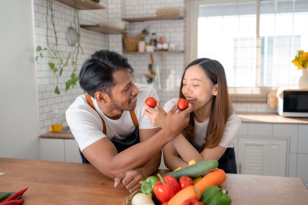 A man and woman in a bright kitchen smile and playfully hold Cocktail Tomatoes near each other’s faces. A variety of fresh vegetables sits on the counter in front of them. GREENHOUSE Goodness