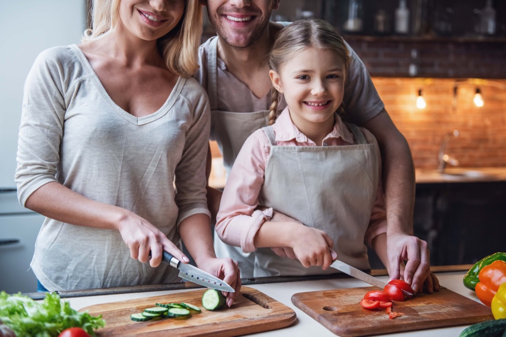 A smiling family of three, two adults and a young girl, prepare vegetables together in a kitchen. Wearing aprons, they slice cucumbers and Cocktail Tomtato tomatoes on wooden boards. GREENHOUSE Goodness