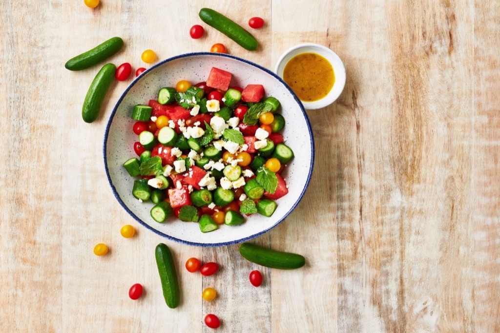 A bowl of colorful salad with diced watermelon, cucumber, cherry tomatoes, feta cheese, and mint leaves, surrounded by mini cucumbers, scattered tomatoes, and a small dish of dressing on a wooden surface. GREENHOUSE Goodness