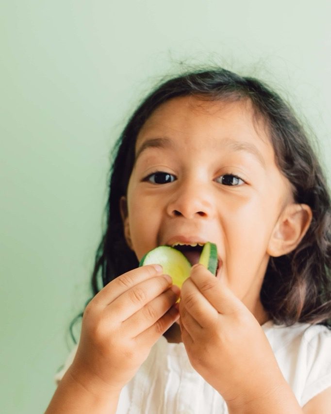 A young child with long dark hair holds and bites into cucumber slices, smiling and wearing a white shirt, against a light green background. GREENHOUSE Goodness
